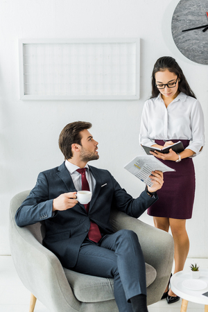 asian businesswoman writing in notebook while businessman sitting on armchair with newspaper and coffee in officeの写真素材
