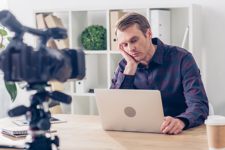 tired handsome male video blogger recording vlog and looking at laptop in officeの写真素材