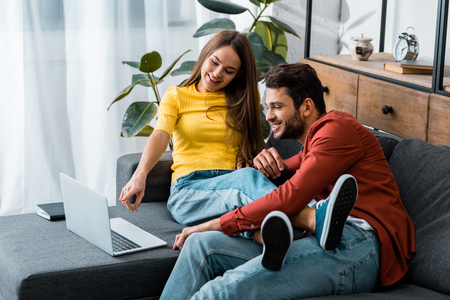 young couple sitting on sofa while girl pointing with finger at laptopの写真素材
