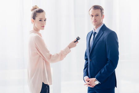 handsome businessman giving interview to attractive journalist with voice recorder in office, looking at cameraの写真素材