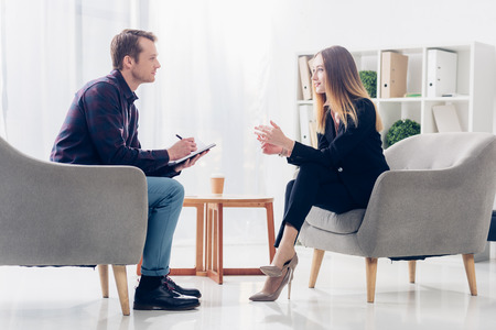 side view of cheerful businesswoman in suit sitting on armchair and giving interview to journalist in officeの写真素材