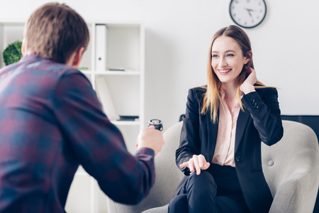 smiling businesswoman in suit touching hair while giving interview to journalist in officeの写真素材