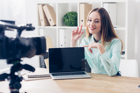 smiling attractive female video blogger recording vlog in office, laptop with blank screen on...
