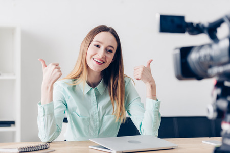 smiling attractive female video blogger recording vlog and showing thumbs up in officeの写真素材