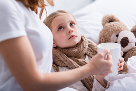 cropped image of mother taking care of sick daughter in bedroom and giving her cup of teaの写真素材