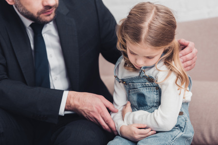 cropped image of father in suit taking care of sick daughter on sofa in living room, she having stomach painの写真素材