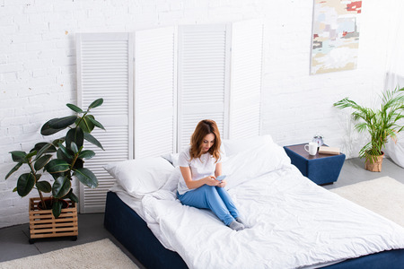 high angle view of woman with ginger hair resting in bed and using smartphone at home on weekend