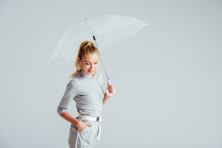 smiling woman in grey clothes and hand in pocket posing with transparent umbrella isolated on greyの写真素材