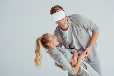 man holding smiling woman in embrace while having virtual reality experience isolated on greyの写真素材