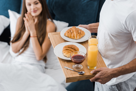 cropped view of man holding wooden tray with breakfast while girl sitting in bedの写真素材