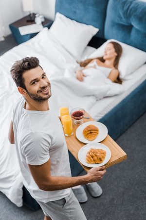 overhead view of smiling man holding wooden tray with breakfast while woman sleeping in bedの写真素材
