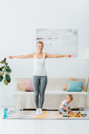 smiling woman exercising with dumbbells and looking at camera and toddler boy playing with multicolored cubes on carpet at homeの写真素材