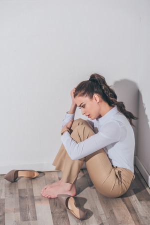 sad woman in white blouse and beige pants sitting on floor near wall at home, grieving disorder conceptの写真素材