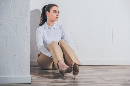 upset woman in white blouse and beige pants sitting on floor near white wall at home, grieving disorder conceptの写真素材