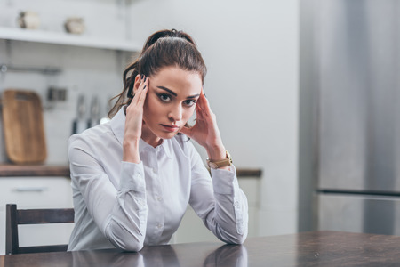 upset woman in white blouse sitting at table and putting hands forehead in kitchen, grieving disorder conceptの写真素材