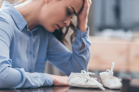 selective focus of sad woman in blue blouse sitting and looking at white baby shoes, grieving disorder conceptの写真素材