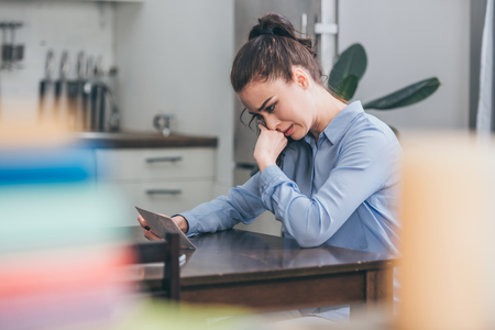 sad woman in blue blouse sitting at wooden table, looking at photo and crying in  kitchen, grieving disorder conceptの写真素材