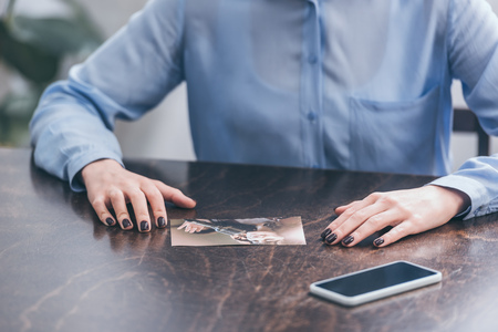 cropped view of woman in blue blouse sitting at wooden table with smartphone and photo at home, grieving disorder conceptの写真素材