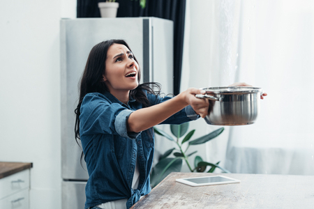 Worried woman in denim short with pot dealing with water damage in kitchenの写真素材