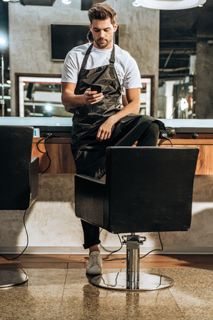 handsome young hairstylist in apron using smartphone in beauty salonの写真素材
