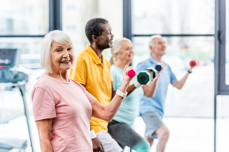 selective focus of smiling senior sportswoman and her friends exercising with dumbbells at sports hallの写真素材