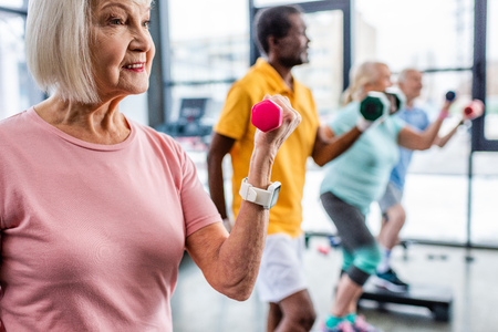 selective focus of senior sportswoman and her friends exercising with dumbbells at gymの写真素材