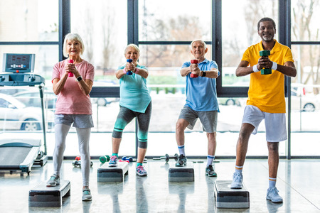 happy senior multicultural sportspeople synchronous exercising with dumbbells on step platforms at gymの写真素材