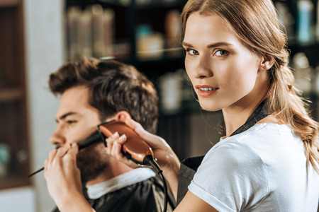 attractive hairstylist looking at camera while trimming hair to male client in beauty salonの写真素材