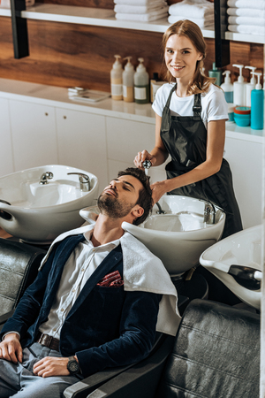 high angle view of hairstylist smiling at camera while washing hair to handsome young man in beauty salonの写真素材