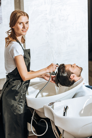 hairstylist smiling at camera while washing hair to handsome young male client in beauty salonの写真素材