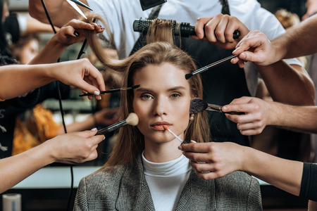 attractive young woman looking at camera while stylists applying makeup and doing hairstyleの写真素材
