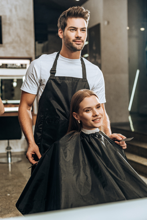 young hairdresser holding comb with scissors and looking at mirror with beautiful woman in beauty salonの写真素材