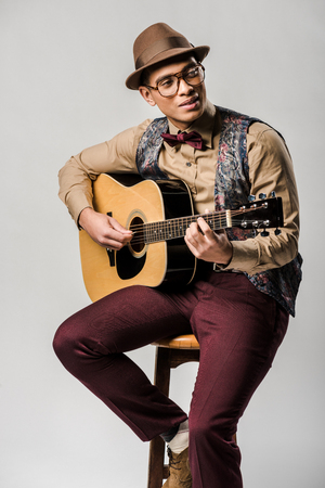 stylish mixed race male musician in hat and eyeglasses playing on acoustic guitar while sitting on chair on greyの写真素材