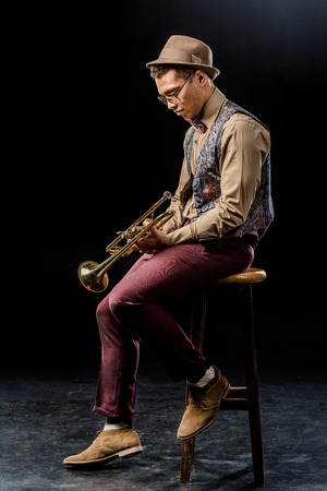 side view of mixed race male musician posing with trumpet while sitting on chair on blackの写真素材