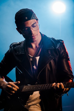 focused male rock star in leather jacket performing on electric guitar on stage with smoke and...