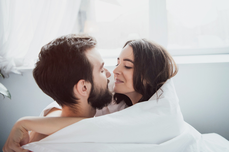 beautiful young happy couple covered in blanket embracing and looking at each other at homeの写真素材