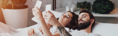 beautiful young couple lying in bed and using smartphonesの写真素材