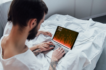 back view of couple lying in bed, holding hands and using laptop with online trade website on screen