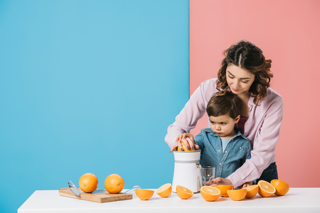 mother with little son squeezing orange juice on white kitchen table on bicolor backgroundの写真素材