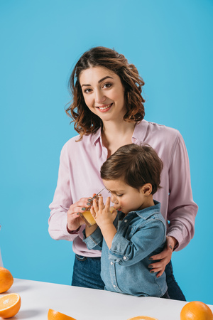 smiling mother helping cute little son in holding glass of fresh orange juice isolated on blueの写真素材