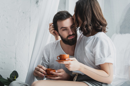 beautiful young couple kissing while having coffee at home in morningの写真素材