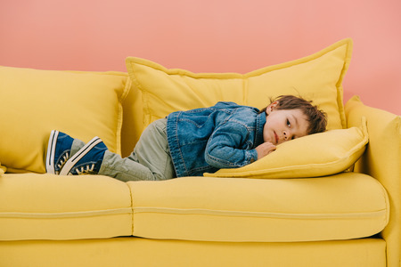 cute little boy in blue cotton jacket and green jeans laying on yellow sofaの写真素材
