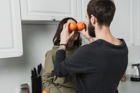 back view of man holding oranges in front of face of woman in kitchen
