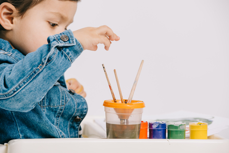 cute little boy choosing painting brush while sitting on highchair with watercolor paints on table isolated on whiteの写真素材