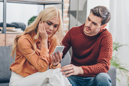 husband looking at upset wife using smartphone on couch, distrust conceptの写真素材