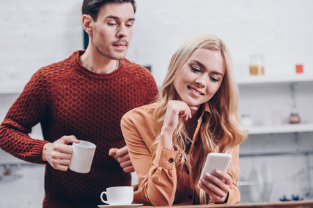 jealous young man holding cup and looking at smiling girlfriend using smartphone in kitchen, mistrust conceptの写真素材
