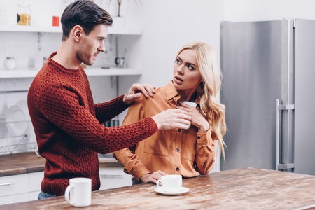 jealous young man looking at scared girlfriend using smartphone in kitchen