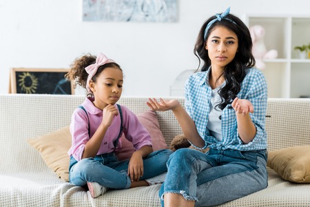 serious african american mother and daughter sitting on sofa at homeの写真素材