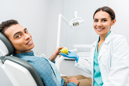 cheerful dentist giving apple to african american patientの写真素材