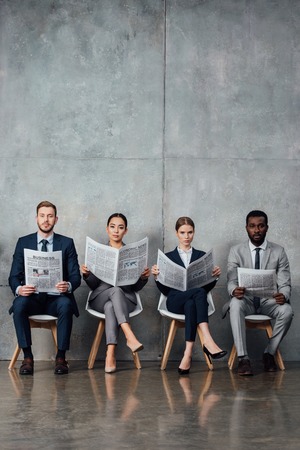 serious multiethnic businesspeople sitting on chairs and reading newspapers in waiting hallの写真素材